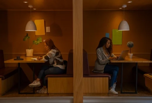 Two people working independently in warmly lit, semi-private booths, one writing in a notebook and the other using a laptop.